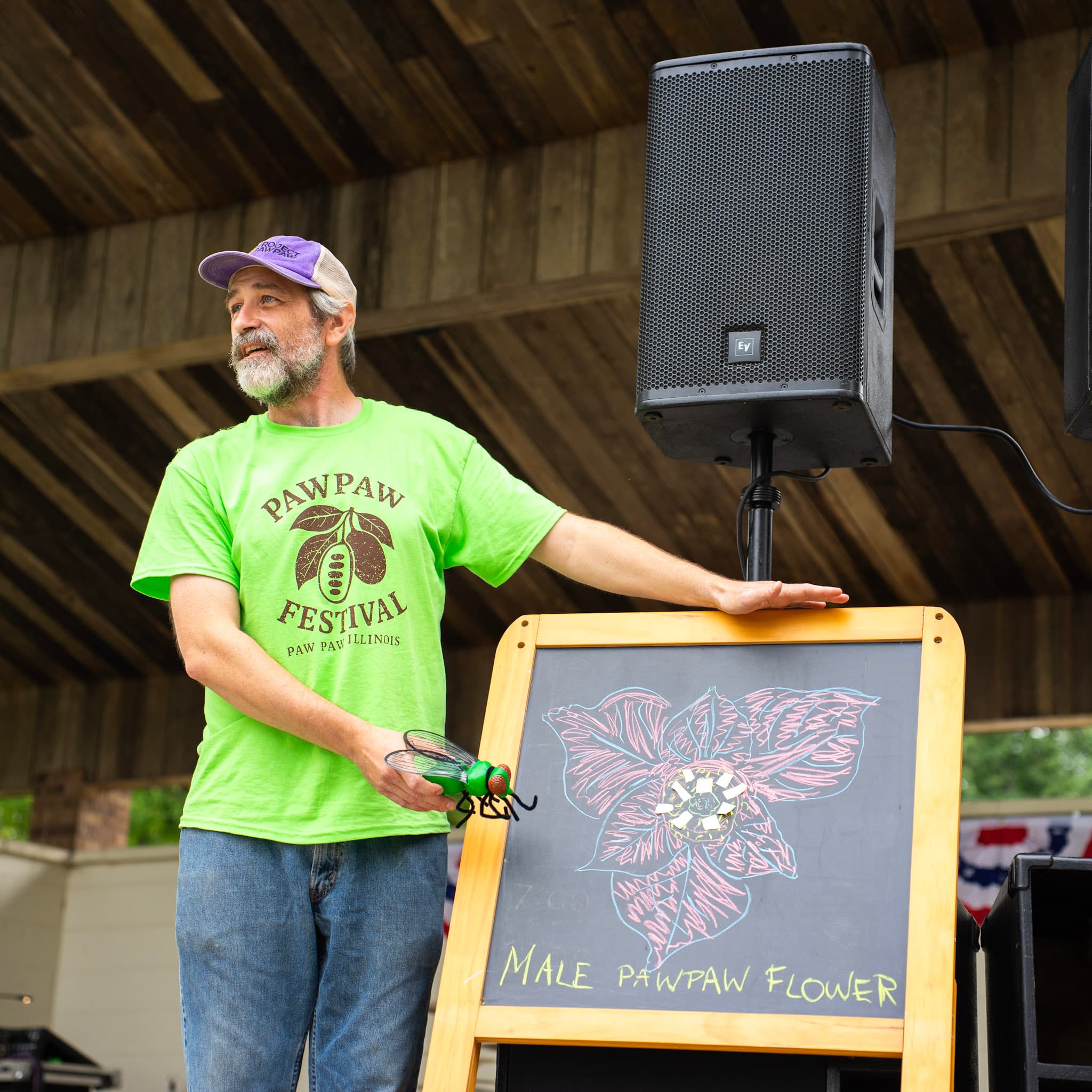Austin Cliffe presenting at the Pawpaw Festival in Paw Paw, Illinois, beside a chalk illustration of a male pawpaw flower